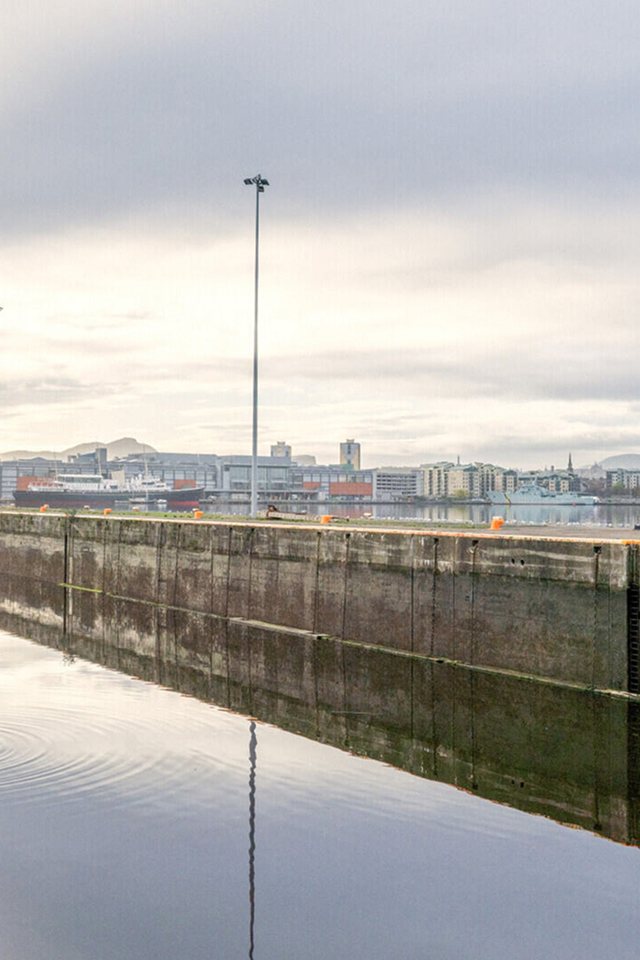 Tall meteorological mast with weather equipment on concrete harbor wall, urban waterfront in background under cloudy sky
