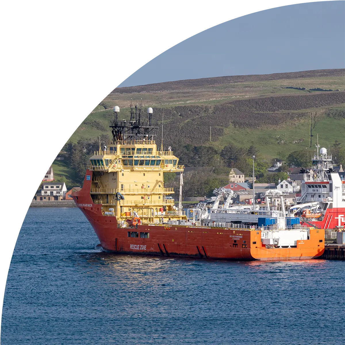 Orange and yellow offshore support vessel docked in harbor with coastal hills and buildings in background