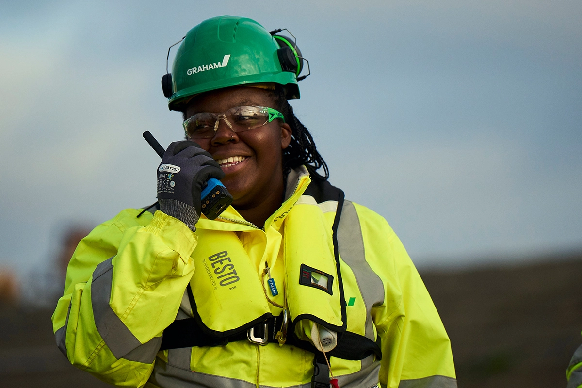 Female offshore wind technician in green safety helmet and yellow high-visibility jacket using radio