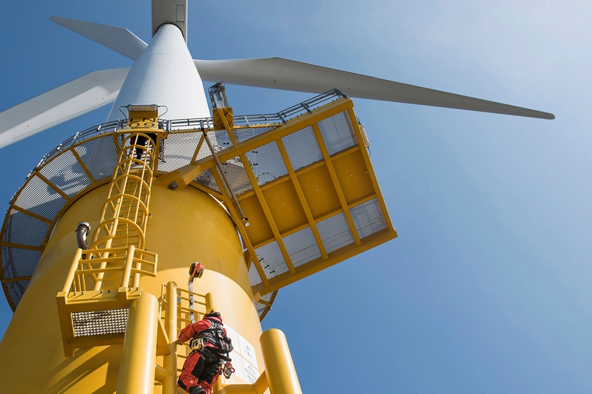 Technician in red safety gear climbing yellow ladder on offshore wind turbine tower under clear blue sky