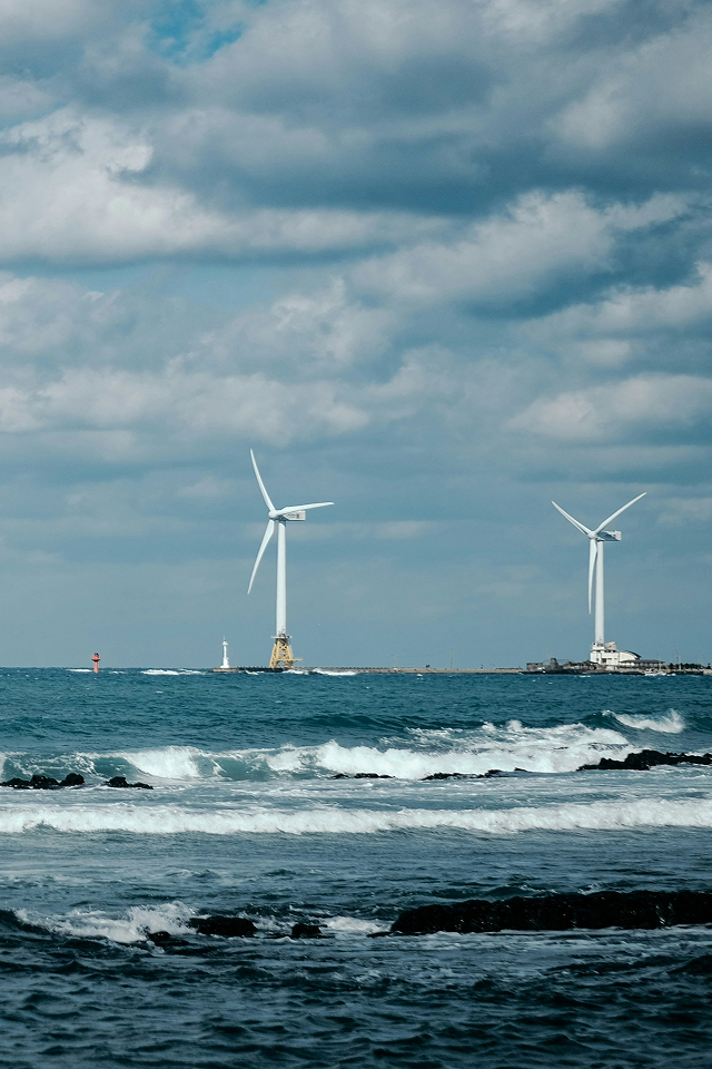 Two offshore wind turbines standing in the ocean with waves and a partly cloudy sky